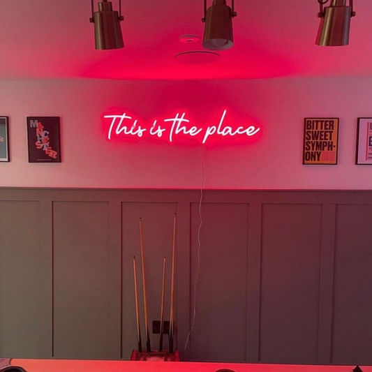 A red neon sign reading 'This is the place' mounted on a wall in a room with dark wood paneling, pendant lights, framed wall art, and pool cues visible below.