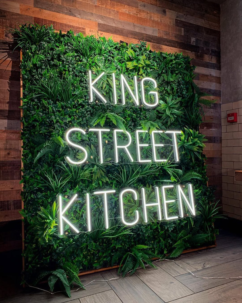 Neon sign reading 'King Street Kitchen' surrounded by green foliage on a wooden wall