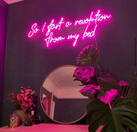 Bright Pink neon sign on a bedroom wall saying “so I start a revolution from my bed” in a handwritten script font.