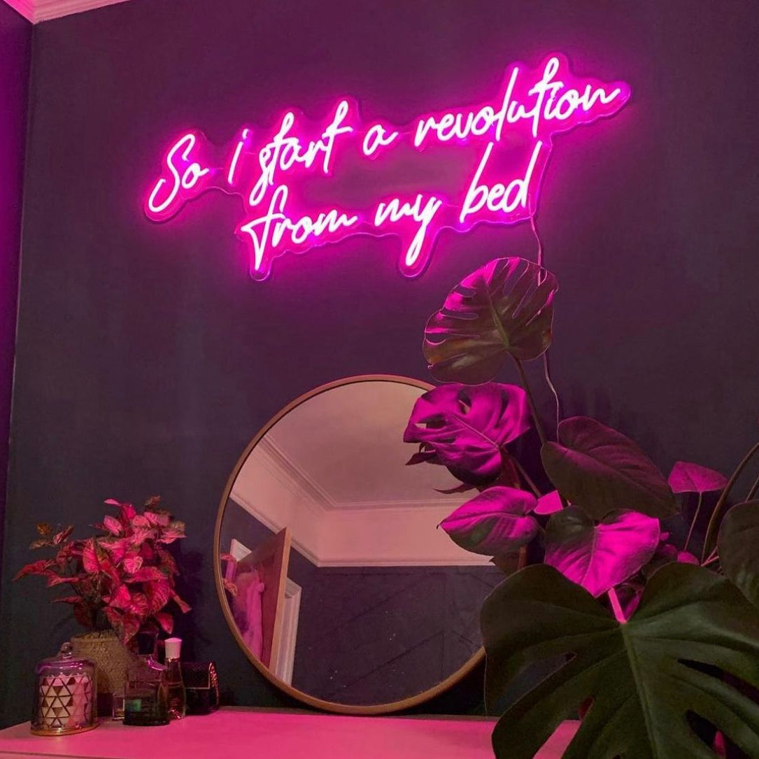 Bright Pink neon sign on a bedroom wall saying “so I start a revolution from my bed” in a handwritten script font.